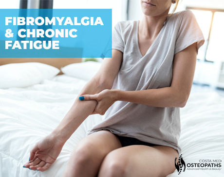 Woman sitting on bed holding her arm, showing signs of pain and fatigue associated with fibromyalgia