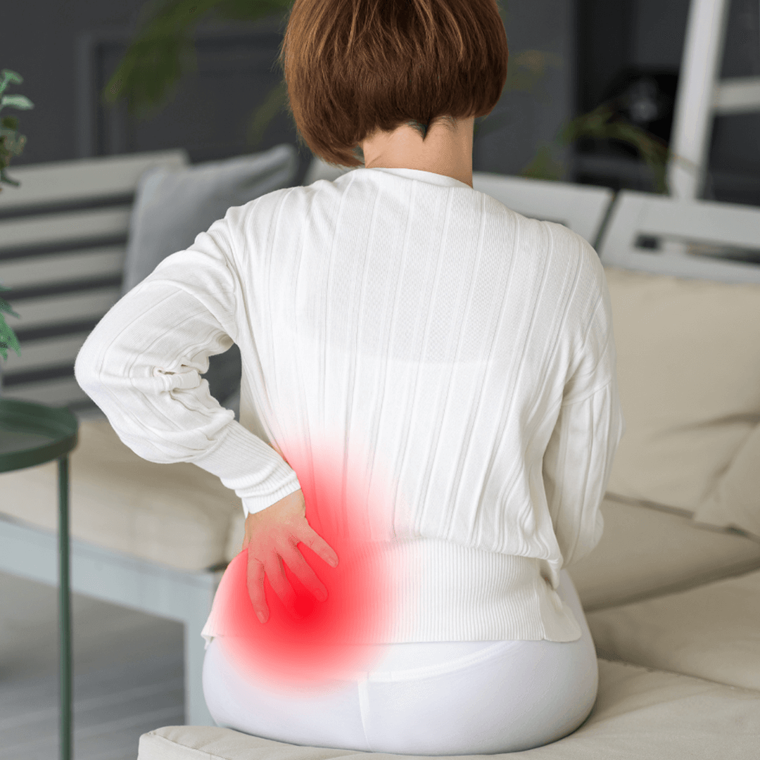 Woman sitting on sofa with lower back pain, highlighted by a red glow over her lumbar spine and sacrum.