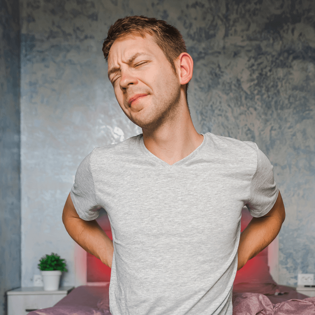 Young man waking up with lower back pain, standing beside bed with red glow highlighting discomfort.