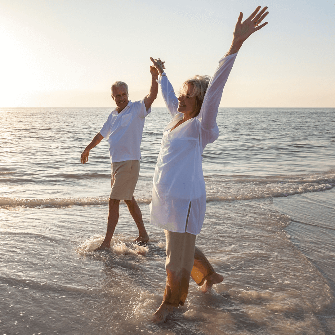 Happy senior couple walking barefoot in the sea at sunset, arms raised in joy.