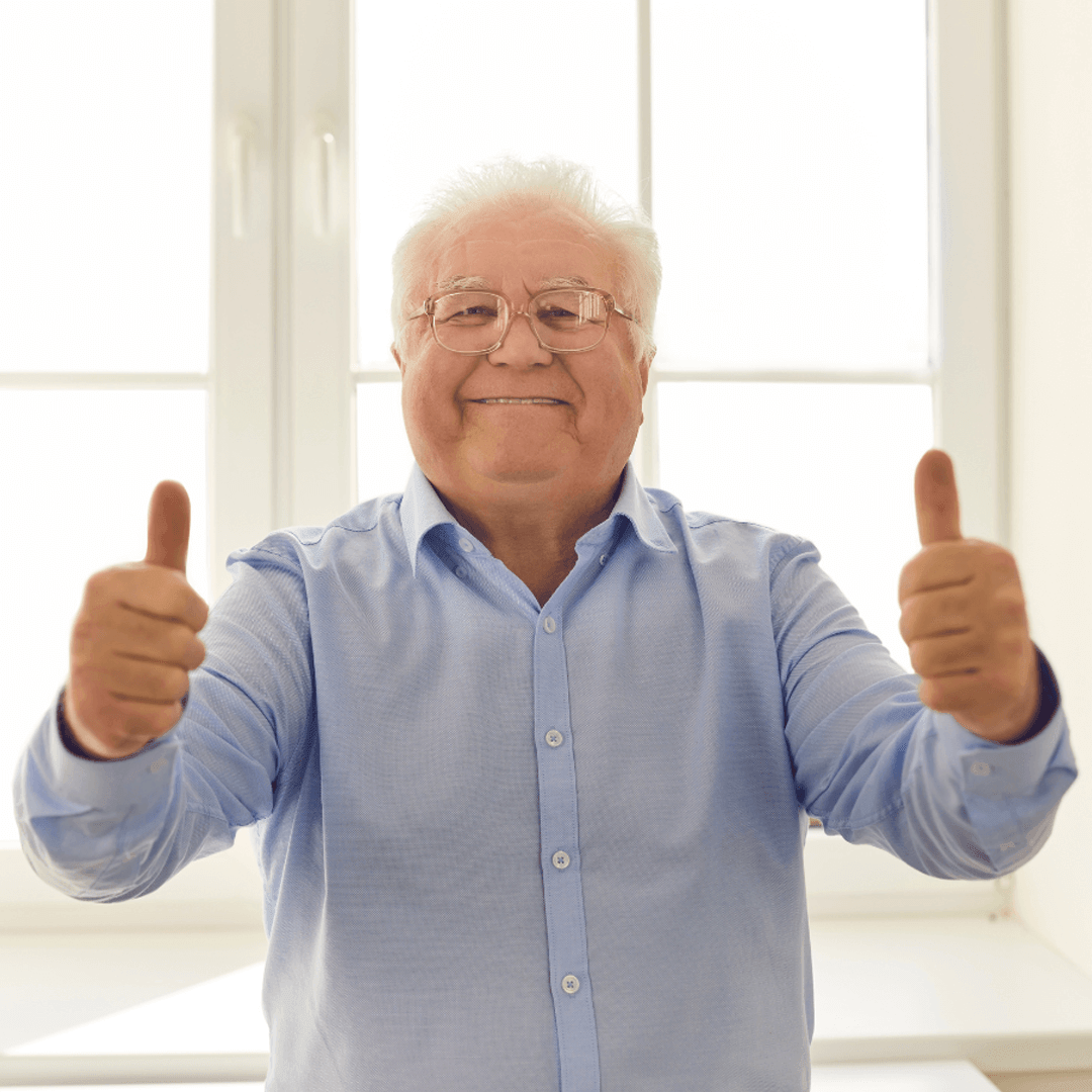 Smiling older man in glasses giving two thumbs up in front of a bright window.